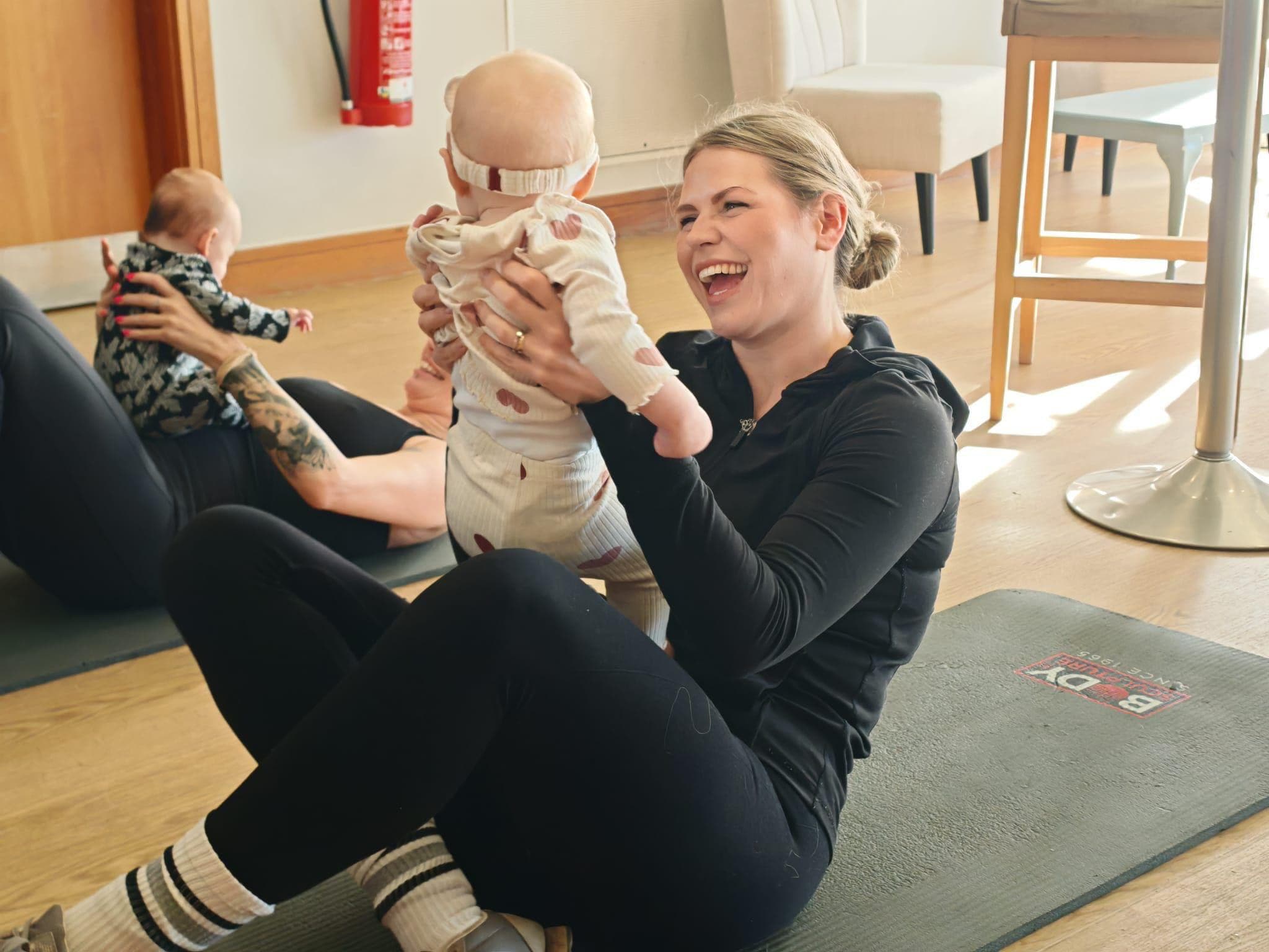 A mum laughing as she lifts her baby above her, on a mat in a community hall, during a Startstrong Fitness CIC class