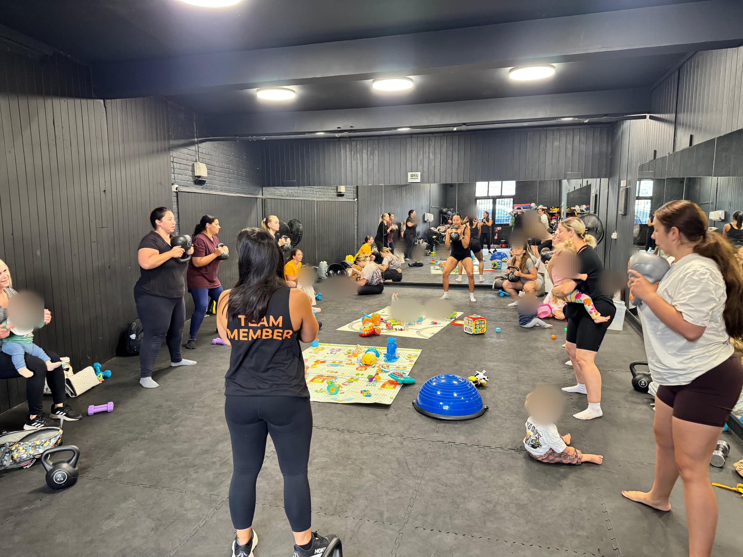A full parent and baby class at Evolution Gym in Stowmarket: parents holding kettlebells in a circle around play mats covered in toys, with babies on the floor