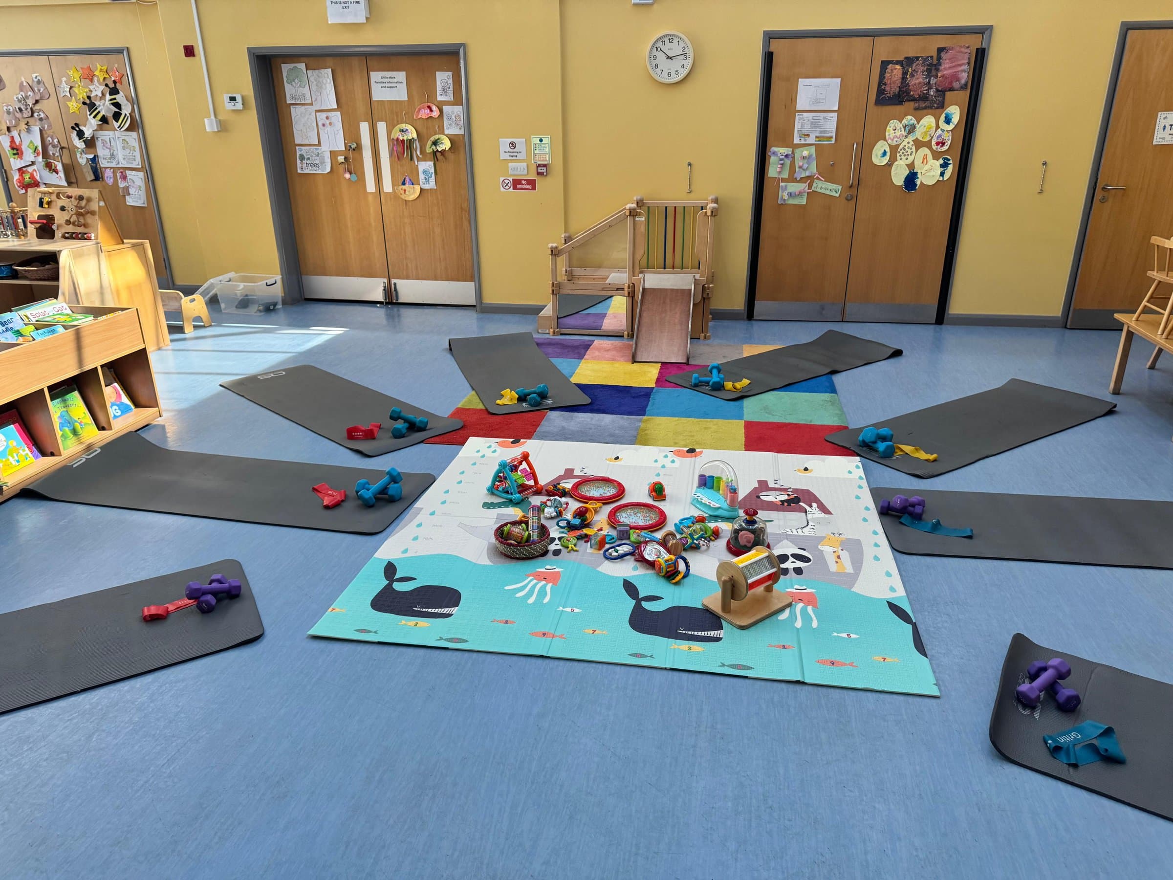 A bright hall with exercise mats laid out in a star around a play mat of toys, ready for class